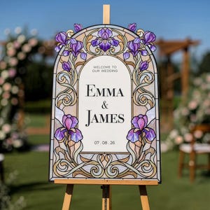 May include: An arched wedding welcome sign featuring a stained glass style border with purple iris flowers and intricate scrollwork. The center white panel displays the text: Welcome to our wedding, Emma & James, and the date 07.08.26.