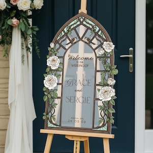 May include: An arched wedding welcome sign featuring a stained glass window design with white roses, green leaves, and two small birds. The text reads: Welcome to the wedding of Grace and Sergie, Boston, DDA, 24 05 06, printed on a clear acrylic panel.