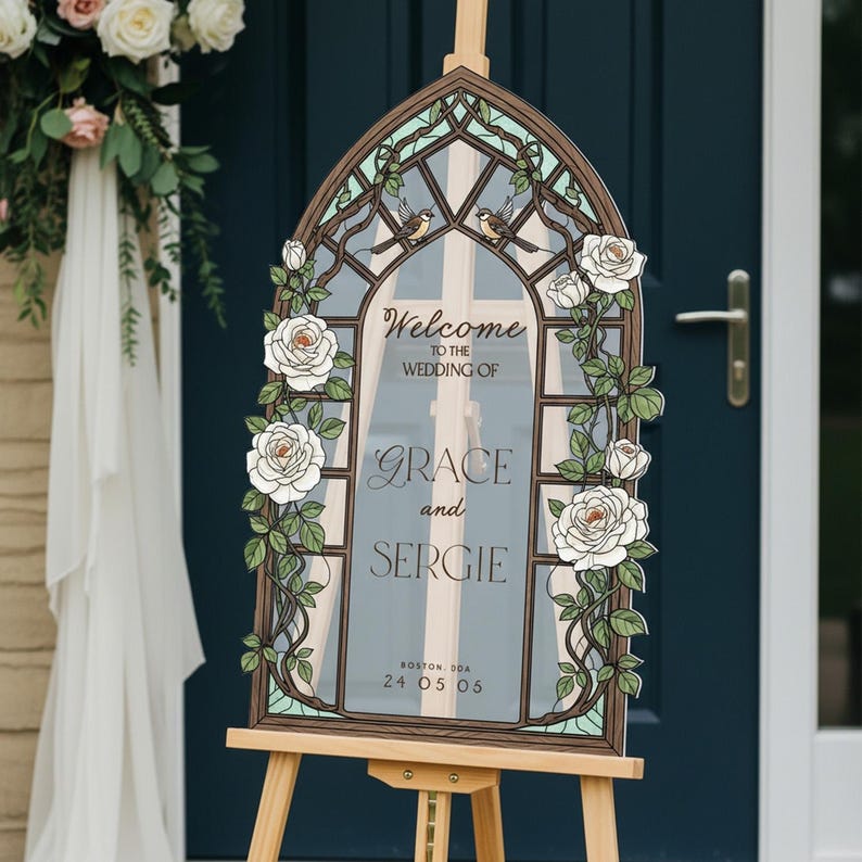 May include: An arched wedding welcome sign featuring a stained glass window design with white roses, green leaves, and two small birds. The text reads: Welcome to the wedding of Grace and Sergie, Boston, DDA, 24 05 06, printed on a clear acrylic panel.
