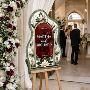 May include: A wedding welcome sign with a deep red center panel featuring the text FOREVER MARTHA and RICHARD 10.01.26. The sign has an arched top and is framed by a decorative border with green and white stained glass style floral patterns.