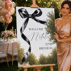 May include: A clear acrylic welcome sign on a wooden easel featuring a large black ribbon graphic. The text reads: WELCOME TO Natalie's 32ND BIRTHDAY 07.15.2026. The sign has a reflective surface showing a blurred outdoor garden setting.
