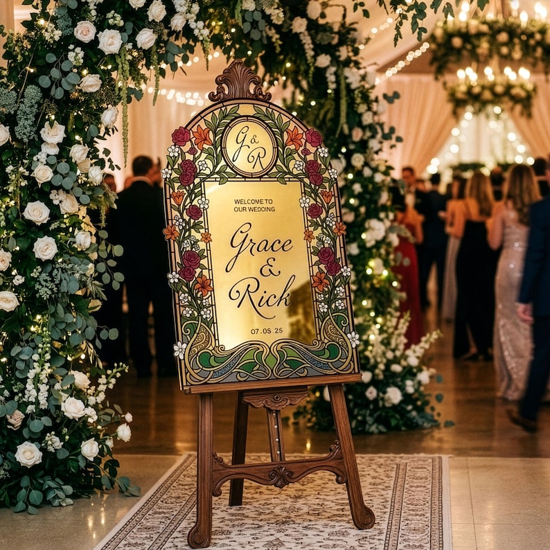 K&ouml;nnte beinhalten: Eine Holzstaffelei h&auml;lt ein dekoratives Schild mit den Namen "Grace & Rick" und dem Hochzeitsdatum. Das Schild hat eine goldene Spiegelfl&auml;che, umgeben von floralen und Jugendstil-Designs. Das Schild befindet sich in einem Hochzeitsort.