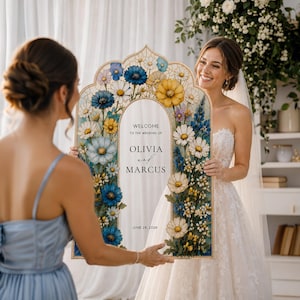 May include: An arched wedding welcome sign featuring a floral border of blue, yellow, and white flowers. The center panel displays the text: WELCOME TO THE WEDDING OF OLIVIA and MARCUS JUNE 24, 2026. The sign has a light-colored wooden frame.