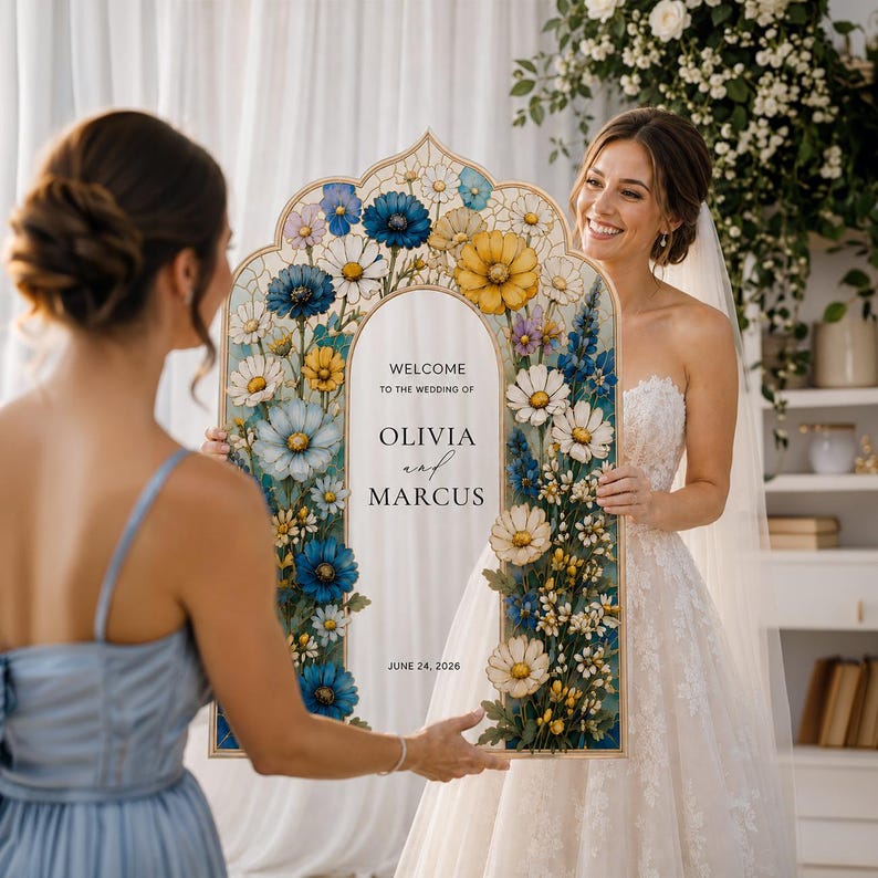 May include: An arched wedding welcome sign featuring a floral border of blue, yellow, and white flowers. The center panel displays the text: WELCOME TO THE WEDDING OF OLIVIA and MARCUS JUNE 24, 2026. The sign has a light-colored wooden frame.
