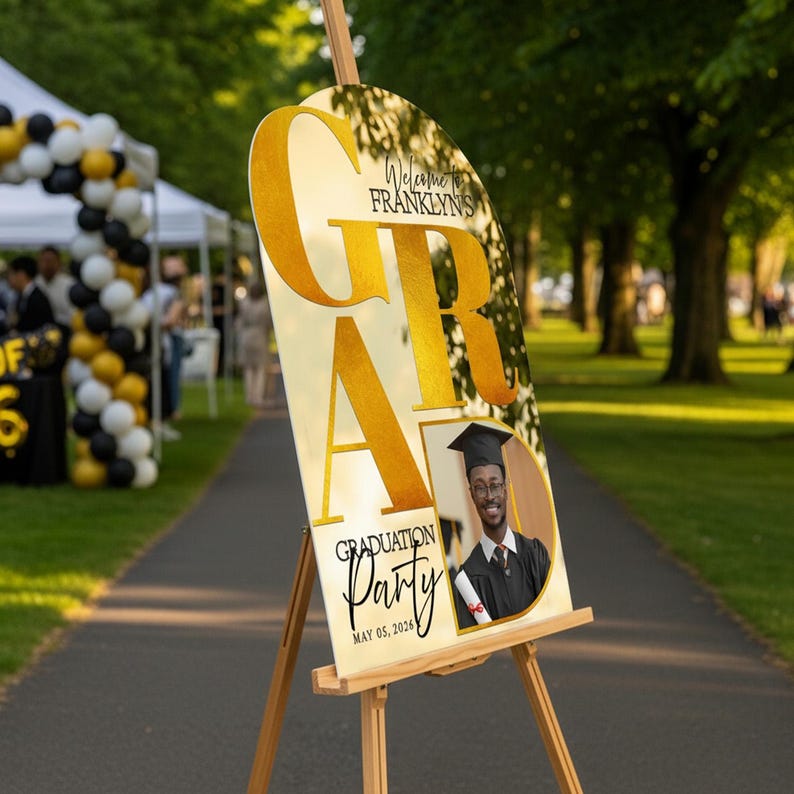 May include: A graduation party sign on a wooden easel. The sign features the word "GRAD" in large gold letters, with the name "Franklyn's" above. A photo of a graduate is included, along with the text "Graduation Party" and the date "May 05, 2026."