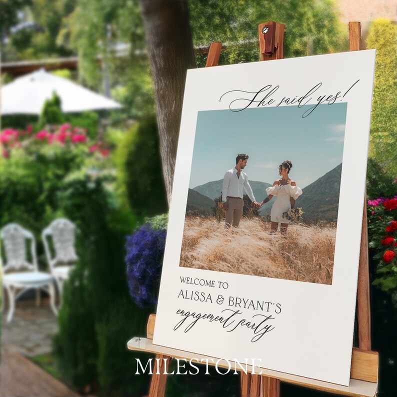 May include: A rectangular white engagement party sign on a wooden easel featuring a central photograph of a couple in a field. The text reads She said yes! at the top and WELCOME TO ALISSA & BRYANT'S engagement party at the bottom in black font.