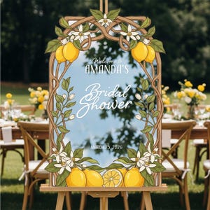 May include: A decorative wooden framed sign on an easel, featuring a mirror with the text "Welcome to AMANDA'S Bridal Shower" and the date "MARCH 25, 2026". The frame is adorned with lemons, white flowers, and green leaves.