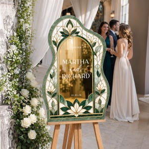 May include: An arched wedding sign with a green and white stained glass pattern border. The center features a gold reflective surface with the text "FOREVER MARTHA and RICHARD 10.01.26" printed in white. The sign is displayed on a wooden tripod easel.
