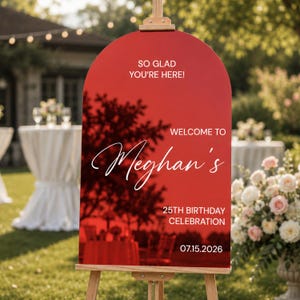 May include: A red arched acrylic welcome sign mounted on a wooden easel. The sign features white text that reads: SO GLAD YOU'RE HERE! WELCOME TO Meghan's 25TH BIRTHDAY CELEBRATION 07.15.2026. The surface is reflective, showing a dark tree silhouette.