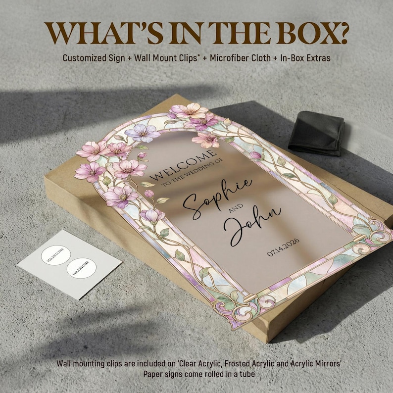 May include: A rectangular wedding sign featuring a stained glass-style floral border in pink and green tones. The text reads: WELCOME TO THE WEDDING OF Sophie AND John 07.14.2026. The sign is placed on a cardboard box with a small black pouch and a white card.