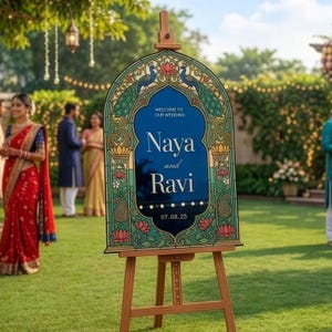 K&ouml;nnte beinhalten: Ein bogenf&ouml;rmiges Hochzeits-Willkommensschild auf einer Holzstaffelei mit tiefblauem Zentrum und dem Text Welcome to our wedding, Naya and Ravi, sowie 07.08.25. Der Rand zeigt kunstvolle Pfauen- und Lotusbl&uuml;tenmotive in Gr&uuml;n, Gold und Rosa.