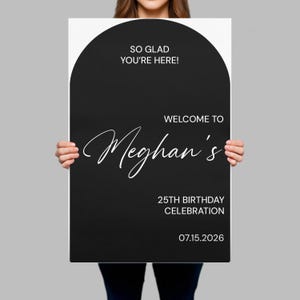 May include: A rectangular black sign with an arched top featuring white text that reads: SO GLAD YOU'RE HERE! WELCOME TO Meghan's 25TH BIRTHDAY CELEBRATION 07.15.2026. The sign has a modern, clean layout with a mix of sans-serif and script typography.