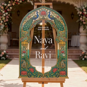 K&ouml;nnte beinhalten: Ein bogenf&ouml;rmiges Hochzeitsschild auf einer Holzstaffelei mit einem Rand im Buntglas-Stil, verziert mit Pfauen- und Lotusbl&uuml;tenmotiven. Der Text lautet: WELCOME TO OUR WEDDING, Naya and Ravi, 07.08.26. Das Design verwendet Blaugr&uuml;n-, Gold- und Rosat&ouml;ne.