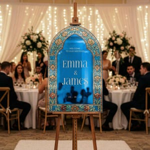 May include: An arched wedding welcome sign on a wooden easel featuring a blue centre panel with white text that reads Welcome to our wedding, Emma & James, 05.16.25. The frame has a decorative stained glass pattern with floral motifs in blue, gold, and red.