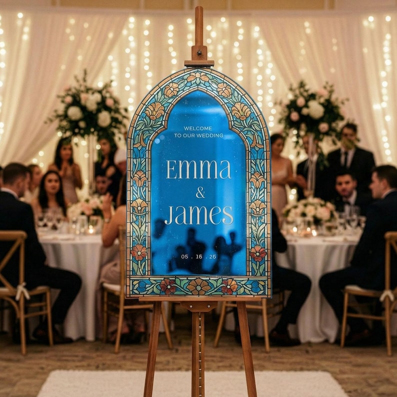 May include: An arched wedding welcome sign on a wooden easel featuring a blue centre panel with white text that reads Welcome to our wedding, Emma & James, 05.16.25. The frame has a decorative stained glass pattern with floral motifs in blue, gold, and red.