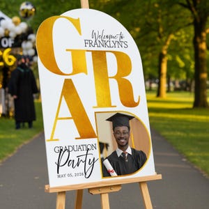 May include: A white graduation party sign on a wooden easel. The sign has gold lettering that says "GRA" and "Graduation Party" with the date May 05, 2026. A photo of a graduate is on the sign.