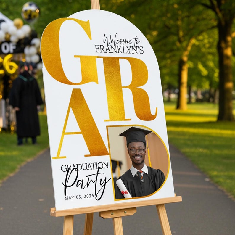 May include: A white graduation party sign on a wooden easel. The sign has gold lettering that says "GRA" and "Graduation Party" with the date May 05, 2026. A photo of a graduate is on the sign.