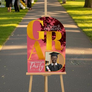 May include: A graduation party sign on a wooden easel. The sign has a pink background with gold lettering that spells out "GRAD". The sign includes the text "Welcome to Franklyn's" and "Graduation Party May 05, 2020". A photo of a graduate is included.