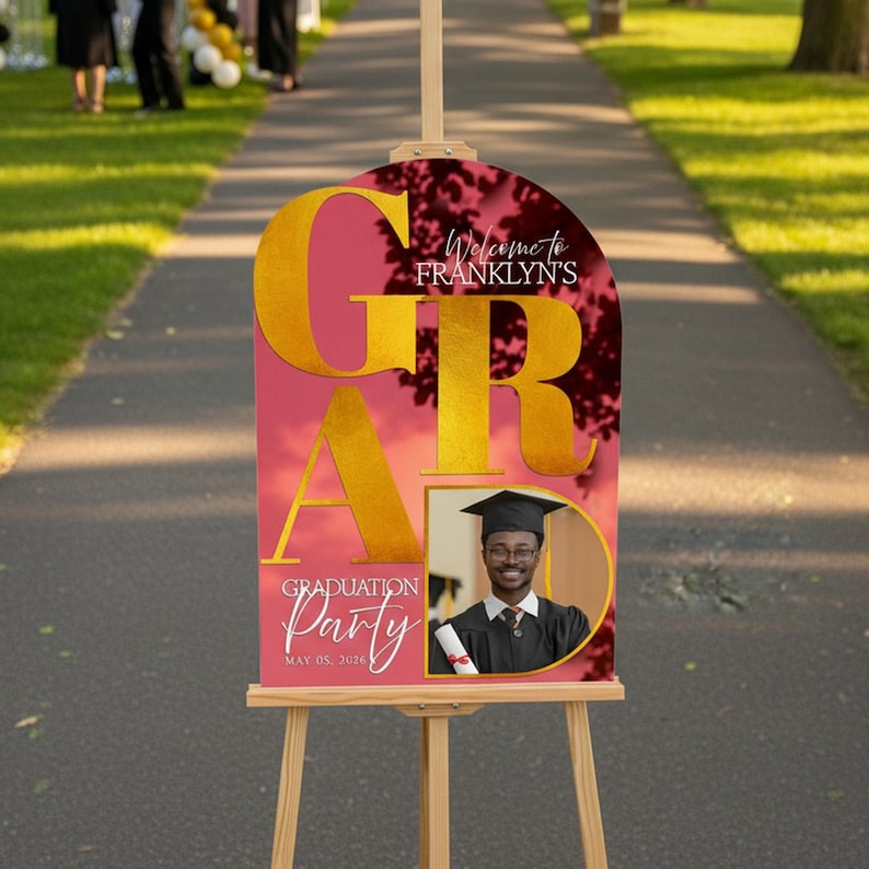 May include: A graduation party sign on a wooden easel. The sign has a pink background with gold lettering that spells out "GRAD". The sign includes the text "Welcome to Franklyn's" and "Graduation Party May 05, 2020". A photo of a graduate is included.
