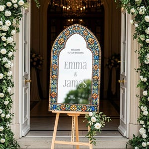 May include: An arched wedding welcome sign featuring a stained glass-style border with blue, orange, and green floral patterns. The centre mirror displays the text: WELCOME TO OUR WEDDING, Emma & James, and the date 05.18.26, resting on a wooden easel.