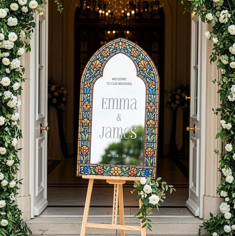 May include: An arched wedding welcome sign featuring a stained glass-style border with blue, orange, and green floral patterns. The centre mirror displays the text: WELCOME TO OUR WEDDING, Emma & James, and the date 05.18.26, resting on a wooden easel.
