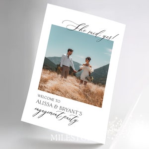May include: A rectangular engagement party sign featuring a central photograph of a couple in a field with mountains in the background. The text reads "She said yes!" at the top and "WELCOME TO ALISSA & BRYANT'S engagement party" at the bottom.