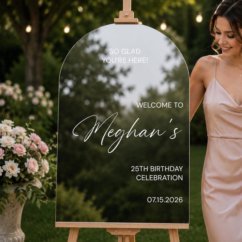 May include: An arched, clear acrylic welcome sign mounted on a wooden easel. The sign features white text reading: SO GLAD YOU'RE HERE! WELCOME TO Meghan's 25TH BIRTHDAY CELEBRATION 07.15.2026. The surface is reflective, showing a blurred outdoor garden.