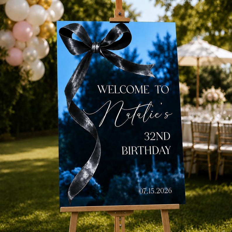 May include: A rectangular birthday welcome sign on a wooden easel featuring a dark blue background with a large black ribbon graphic. The text reads: WELCOME TO Natalie's 32ND BIRTHDAY 07.15.2026. The design includes a deep blue forest scene.
