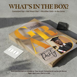May include: A graduation party sign with the word "GRAD" in gold lettering. The sign includes a photo of a graduate and the text "Graduation Party May 05, 2026". The box includes wall mount clips and a microfiber cloth.