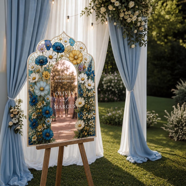 May include: An arched wedding welcome sign on a wooden easel, featuring a mirror center with the text 'WELCOME TO THE WEDDING OF OLIVIA AND MARCUS'. The frame is decorated with a vibrant floral border of blue, yellow, and white daisy-like flowers.