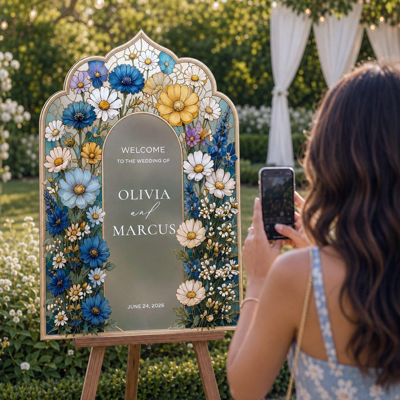 May include: An arched wedding welcome sign featuring a gold frame and a frosted center panel with the text Welcome to the wedding of Olivia and Marcus June 24, 2026. The border is decorated with a colorful illustration of blue, yellow, and white flowers.