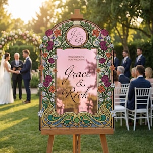K&ouml;nnte beinhalten: Eine Holzstaffelei zeigt ein Hochzeitsschild mit einer rosa verspiegelten Oberfl&auml;che. Das Schild ist mit farbenfrohen Blumen- und Blattmotiven umrahmt und tr&auml;gt die Namen "Grace & Rick" und das Datum "07.08.26". Die Worte "Welcome to our Wedding" sind ebenfalls zu sehen.