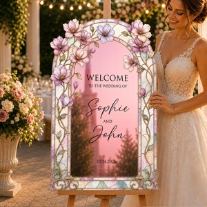 May include: An arched wedding welcome sign featuring a stained glass border with pink and purple floral illustrations. The centre text reads: WELCOME TO THE WEDDING OF Sophie AND John 14.07.2026. The sign is mounted on a wooden easel stand.