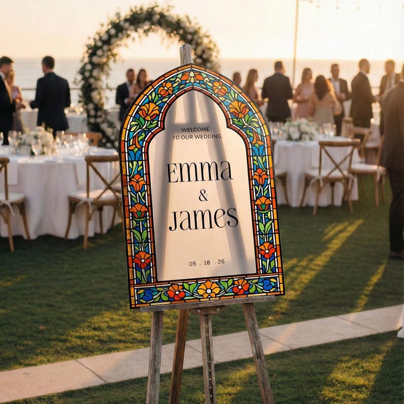 May include: A wedding welcome sign on a wooden easel featuring a stained glass-style border with orange, blue, and green floral patterns. The centre text reads: WELCOME TO OUR WEDDING, EMMA & JAMES, 05 . 18 . 26, printed in black serif font.