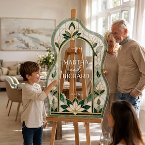 May include: A custom wedding welcome sign featuring a stained glass-inspired floral pattern in shades of green and white with gold outlines. The sign displays the text FOREVER, MARTHA and RICHARD, and the date 10.01.26, mounted on a wooden easel.