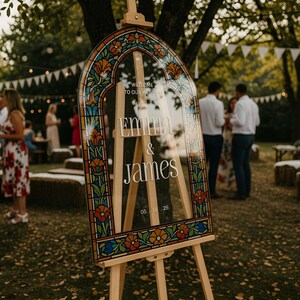 May include: An arched acrylic wedding sign featuring a colourful stained glass border pattern with orange, blue, and green floral motifs. The sign displays the white text: Welcome to our wedding, Emma & James, 05.18.26, mounted on a light wooden easel.