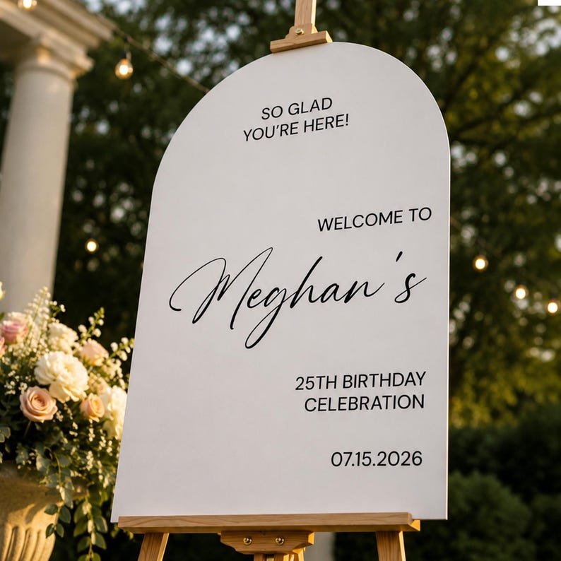May include: A white arched welcome sign on a wooden easel featuring black text that reads SO GLAD YOU'RE HERE! WELCOME TO Meghan's 25TH BIRTHDAY CELEBRATION 07.15.2026. The sign has a smooth surface and is displayed outdoors with a floral arrangement nearby.