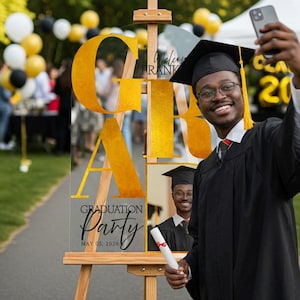 May include: A clear acrylic graduation party sign with gold lettering spelling out "GRAD" and "Party" in script. The sign is on an easel, with a photo of a graduate and the date May 05, 2026. A graduate in a cap and gown takes a selfie.