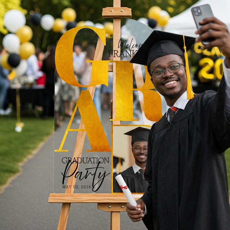 May include: A clear acrylic graduation party sign with gold lettering spelling out "GRAD" and "Party" in script. The sign is on an easel, with a photo of a graduate and the date May 05, 2026. A graduate in a cap and gown takes a selfie.