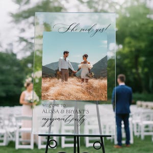 May include: A clear acrylic rectangular sign featuring the text "She said yes!" at the top and "WELCOME TO ALISSA & BRYANT'S engagement party" at the bottom. A square photograph of a couple in a field is centered between the text on the sign.