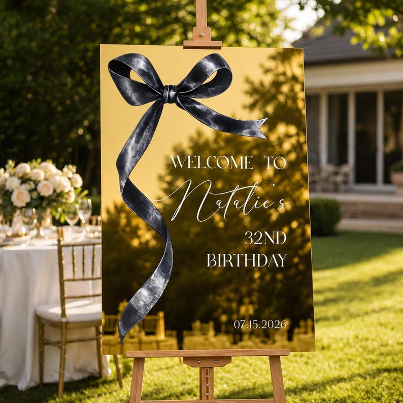 May include: A rectangular gold mirrored welcome sign featuring a large black ribbon graphic. The text reads: WELCOME TO Natalie's 32ND BIRTHDAY 07.15.2026. The sign is mounted on a wooden easel, displaying a reflective surface with a dark bow design.