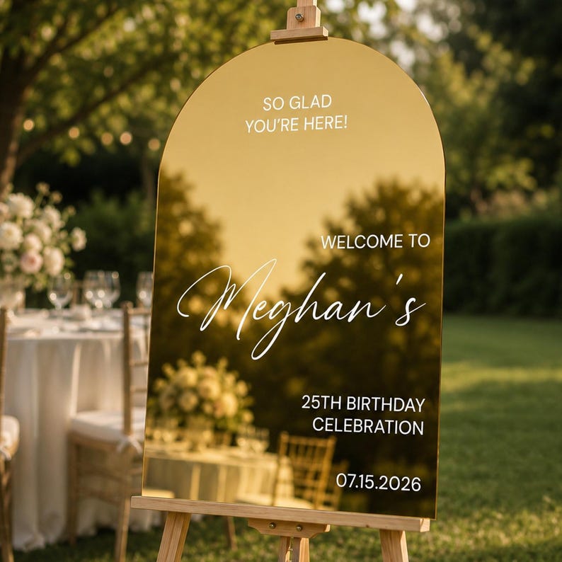May include: An arched gold mirror sign featuring white text that reads: SO GLAD YOU'RE HERE! WELCOME TO Meghan's 25TH BIRTHDAY CELEBRATION 07.15.2026. The sign is mounted on a wooden easel, reflecting the surrounding outdoor environment.