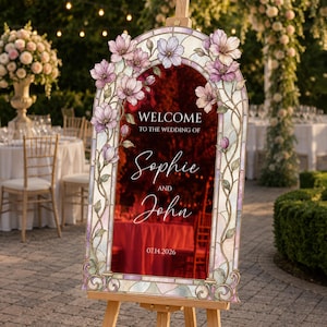 May include: An arched wedding welcome sign featuring a stained glass border with pink and purple floral accents. The centre displays the text: WELCOME TO THE WEDDING OF Sophie AND John 07.14.2026. The sign is mounted on a wooden easel stand.
