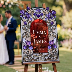 May include: An arched wedding welcome sign featuring a stained glass-style border with purple iris flowers and gold scrollwork. The center is a deep red panel with white text reading: WELCOME TO OUR WEDDING, EMMA & JAMES, 07.08.26.