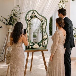 May include: An arched wedding sign with a green stained glass border featuring floral motifs. The center is clear with white text reading MARTHA and RICHARD and the date 10.01.26. The sign is displayed on a wooden tripod easel stand.