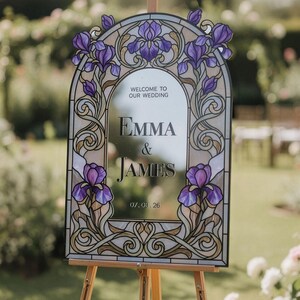 May include: An arched wedding welcome sign featuring a stained glass border with purple iris flowers and gold scrollwork. The center mirror displays the text: WELCOME TO OUR WEDDING, EMMA & JAMES, and 07.03.26 in a dark serif font.