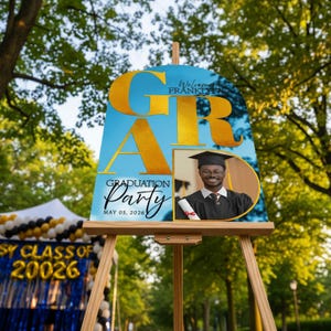 May include: A graduation party sign on a wooden easel. The sign has a blue background with gold lettering that spells "GRAD". A photo of a smiling graduate is on the right. The sign reads "Graduation Party May 05, 2026".