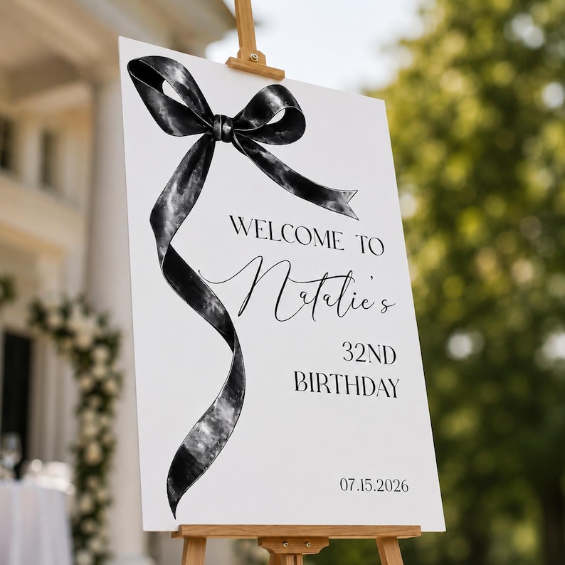 May include: A white rectangular welcome sign on a wooden easel featuring a large black watercolor-style ribbon bow graphic. The text reads: WELCOME TO Natalie's 32ND BIRTHDAY 07.15.2026. The design uses a mix of bold sans-serif and elegant script fonts.