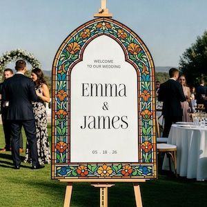 May include: An arched wedding welcome sign on a wooden easel featuring a stained glass-style border with orange, yellow, blue, and green floral patterns. The white centre displays the text: WELCOME TO OUR WEDDING, EMMA & JAMES, 05 . 18 . 26.