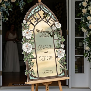 May include: An arched wedding welcome sign featuring a stained glass window design with white roses, green vines, and two small birds. The text reads: Welcome to the wedding of Grace and Sergie, Boston, USA, 24.05.26. The sign is displayed on a wooden easel.
